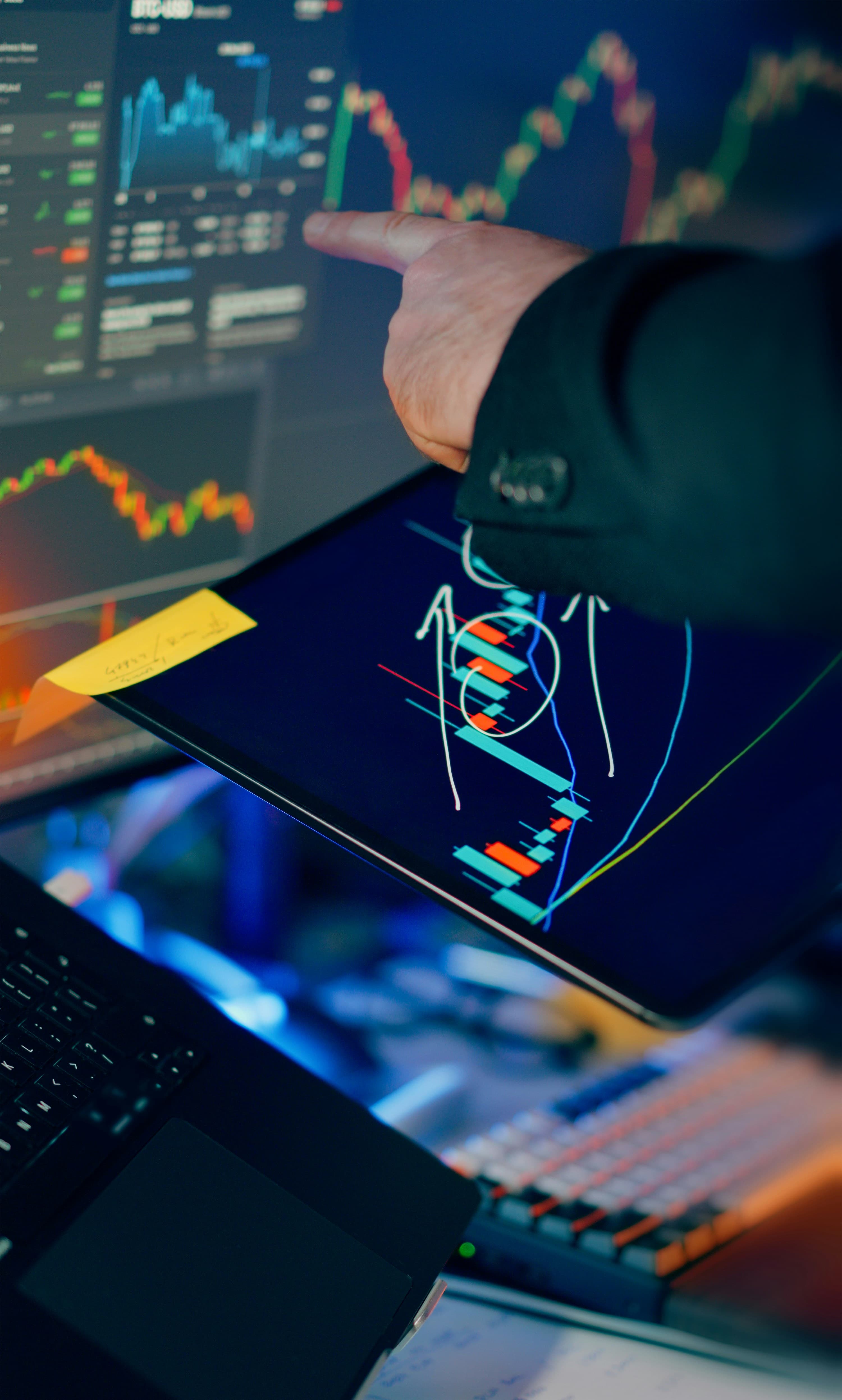 A hand annotating transaction charts across a tablet and monitors in a dark workspace